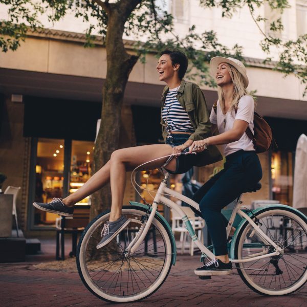 Smiling woman sitting on bicycle handlebar with her friend. Beautiful women enjoying on bicycle in the city.